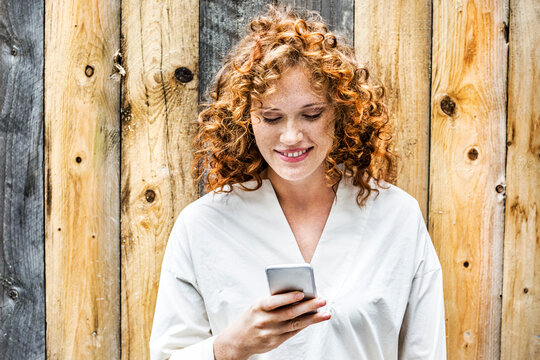 Portrait of smiling young woman looking at cell phone in front of wooden wall