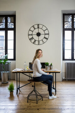 Portrait of smiling woman sitting at desk at home under large wall clock