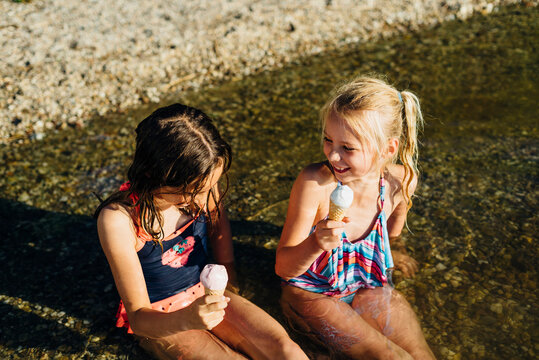 Two girls sitting in water at lakeshore eating icecream