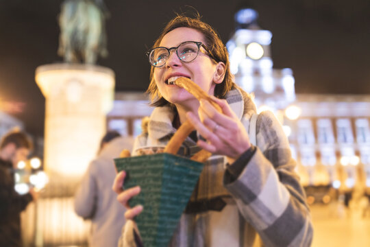 Spain, Madrid, young woman in the city at night eating typical churros with chocolate