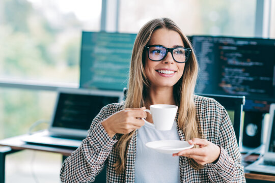 Smiling female developer enjoying a coffee break at her modern office desk with multiple monitors showing code and a relaxed, productive tech work environment