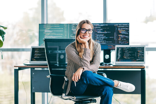 Smiling female software developer sitting in modern office with laptops and multiple monitors showing code, relaxed programmer working remotely and enjoying tech-focused creative workspace