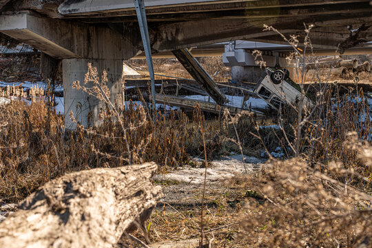 Irpin, Ukraine - 07 March 2026: View of the Irpin Bridge's skeletal remains, a stark testament to destruction, with a vehicle suspended precariously amidst the rubble and withered vegetation.