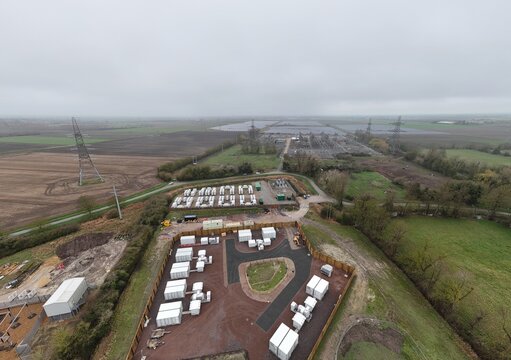 Aerial view of rows of white containers contrasting against the reddish-brown earth, with electrical towers standing tall in the distance, Burwell, United Kingdom.