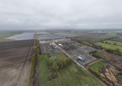 Aerial view of the electrical substation and solar farm, where silver panels meet the grey infrastructure under a vast, muted sky, Burwell, United Kingdom.