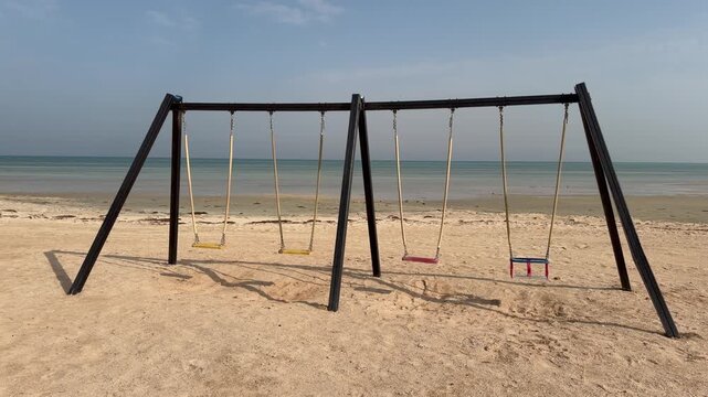 A quiet winter beach in Sila shows wooden swing seats on soft sand, facing gentle waves near the Abu Dhabi&ndash;Saudi border.