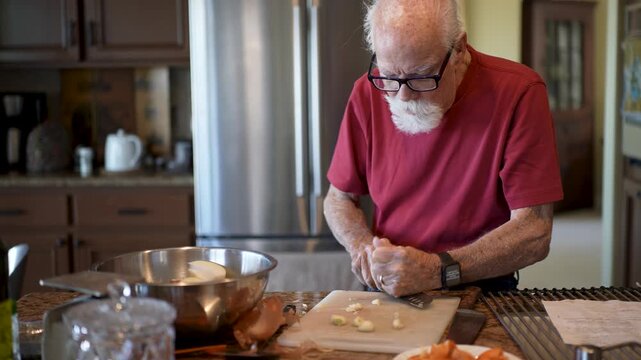 Elderly man spends time in home kitchen mashing and mincing garlic for his cooking preparations.