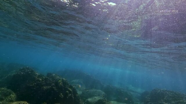 Sunbeams pierce clear blue tropical ocean water, creating a beautiful aquatic display. Light dances on the rocky bottom, revealing natural depth