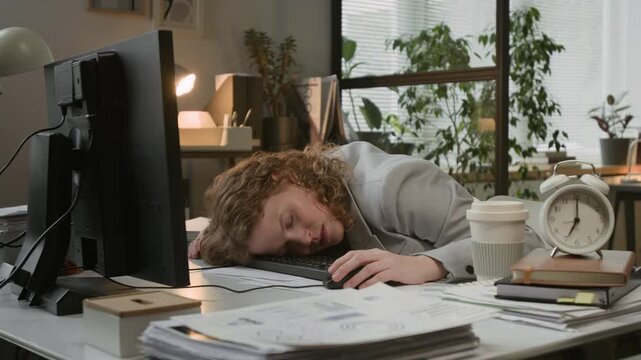 Low angle shot of young exhausted Caucasian woman with curly hair sleeping face down on keyboard with mouse in hand at her office desk