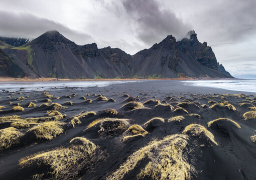 Aerial view of the stark black sand beach meeting jagged, imposing peaks under a sky heavy with dramatic clouds, Stokksnes, Sveitarfelagid Hornafjordur, Iceland.