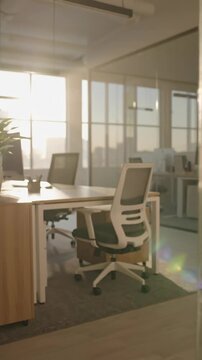 Blurred modern office interior with chair and desk illuminated by warm sunlight through large windows. Defocused workspace background. Vertical video.