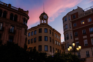 An ornate corner building with a domed turret and clock anchors a Cordoba, Spain street, with curved brick facades, iron balconies, and street lamps glowing at dusk. © True Pixel Art