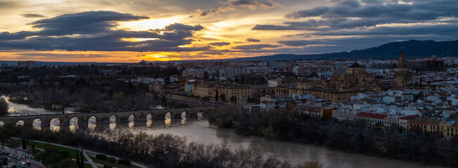 Pedestrians cross Puente Romano as storm clouds part over Cordoba, Spain. The Guadalquivir reflects sunset light near the Mezquita Catedral and whitewashed quarters. © True Pixel Art