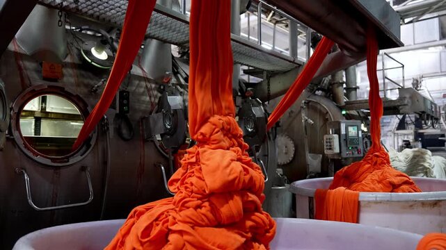 Bright orange fabric flowing from large dyeing machines in a textile factory. Professional industrial footage reflecting technical process, production control and large scale manufacturing.