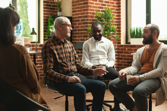 Caucasian men having conversation as black man smiles at camera in brick wall room. Young and old male individuals sitting together and discussing about mental health at aa group meeting.