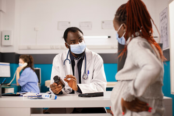 Fototapeta premium Male physician explains prenatal care while holding bottle of pills to pregnant black woman. White female nurse using telephone in background while doctor gives prescribed medicine to expectant mother
