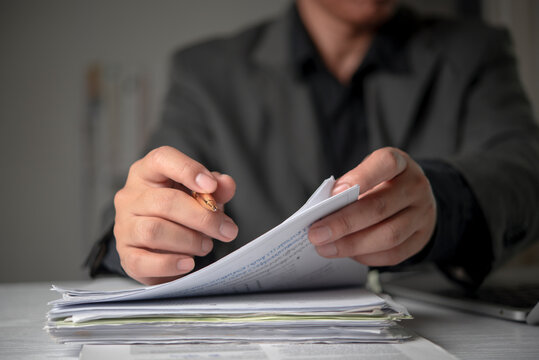 Businessman hands holding gold pen reviewing thick stack paperwork documents office desk. Corporate executive analyzing legal contract financial audit administrative workload concept.
