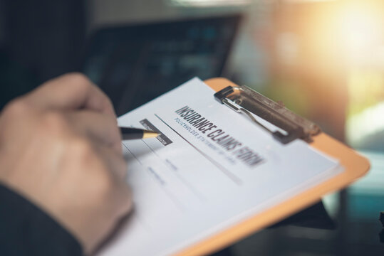 Hand holding pen writing insurance claims form clipboard office desk. Person filling policyholder statement paperwork medical auto health accident property coverage financial protection.