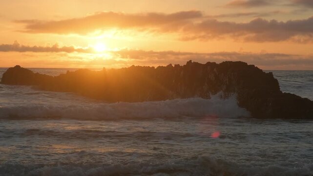 Sandy Ocean shore with rolling waves with an orange sun on the horizon at sunset. Sand beach with rolling waves hitting a big rock. Contemplation of the sea waves during sunset.