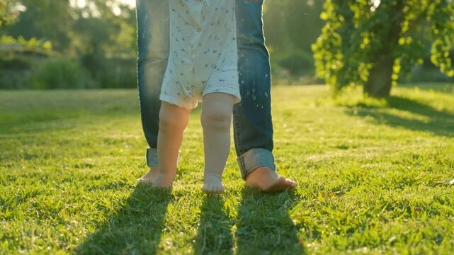 Walking children's bare feet on a green lawn close-up. Child  learns to take the first steps.  Baby learns to walk with the help of his mother on a green grass in the park. Real time 4k video.
