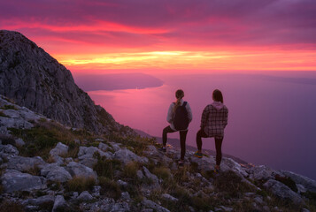 Obraz premium Two women hikers with backpacks standing on rocky mountain peak at sunset, overlooking the sea with colorful pink sky. Outdoor travel adventure. Silhouette of girls on mountain at dusk. Lifestyle