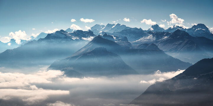 Panoramic aerial view of the Swiss Alps with snow-capped peaks and low clouds at sunrise. Bright morning light and blue sky over rugged alpine mountains in Switzerland at dawn. Top view. Landscape