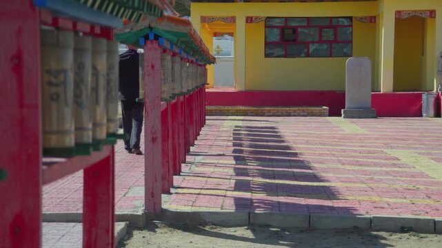 Side View Prayer Wheels Leading Courtyard Asian Monk Walks Past Row Of Brass Wheels Under Yellow Temple Facade, Long Shadow On Patterned Pavement, Rhythmic Ritual, Calm Heritage Atmosphere