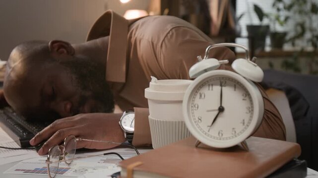 Close-up shot of exhausted young African American man sleeping face down at computer desk with alarm clock in modern office