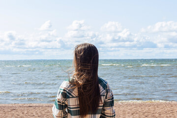 Young woman with long hair standing on the beach, facing the sea, enjoying the view with waves and a serene atmosphere.