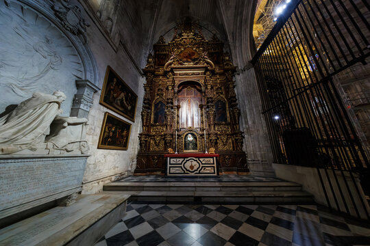 Baroque altarpiece with gilded wood, paintings, and reliquary stands in a side chapel in Seville, Spain, beside a white marble tomb and checkerboard stone floor.