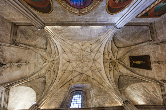 Vaulted ceiling and star ribbed vault inside Seville Cathedral. Gothic columns rise to pointed arches, a tall grilled window and paintings frame a sacred interior.