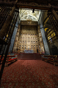 Seville, Spain. A soaring Gothic nave frames the monumental gold retablo of the Capilla Mayor inside Seville Cathedral, with ribbed vaults, stepped altar, and warm lighting.