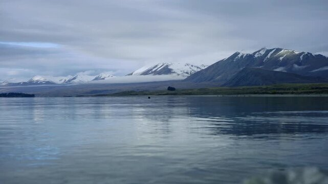 A pastoral and dreamy view of a snow capped mountains and their reflections on a calm water lake at twilight, with some light clouds. a 4K video clip, lake Tekapo, New Zealand.