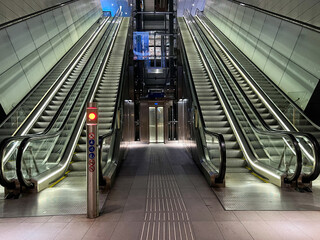 Modern escalators in a well-lit urban subway station with glass walls and an elevator visible in the center, showcasing a sleek architectural design and contemporary infrastructure © Ann