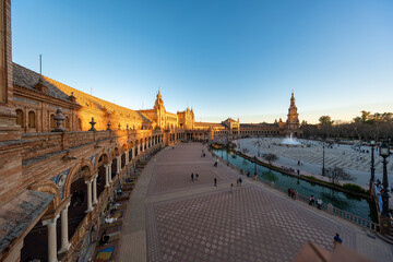 Fototapeta premium Late afternoon Plaza de Espana view with north tower and fountain in Seville, Spain. Wide angle scene shows arches, canal bridges, azulejo benches, and strolling visitors.