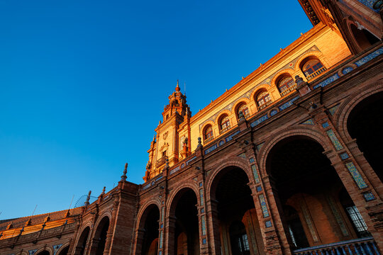 Low angle view of Plaza de Espana in Seville, Spain shows sweeping arcades and a tower, warm late day light, patterned azulejo tiles, and deep shadows across arches.