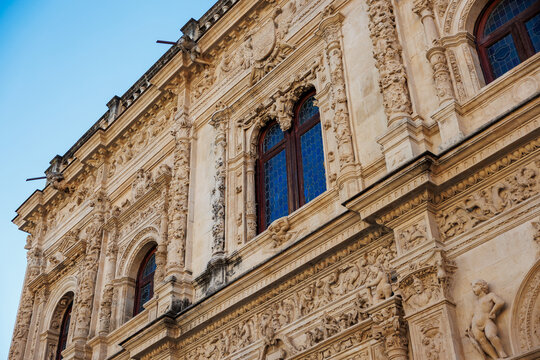 Seville, Spain civic landmark shows Plateresque facade, arched windows with blue leaded glass, sculptural columns, cherubs, grotesques, and crests in warm afternoon light.