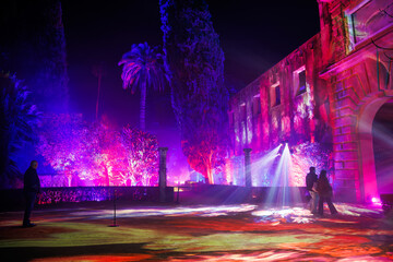 Naklejka premium Night light festival in Real Alcazar gardens, Seville. Magenta, violet, and blue projections wash stone walls as visitors wander past columns and an arched portal.