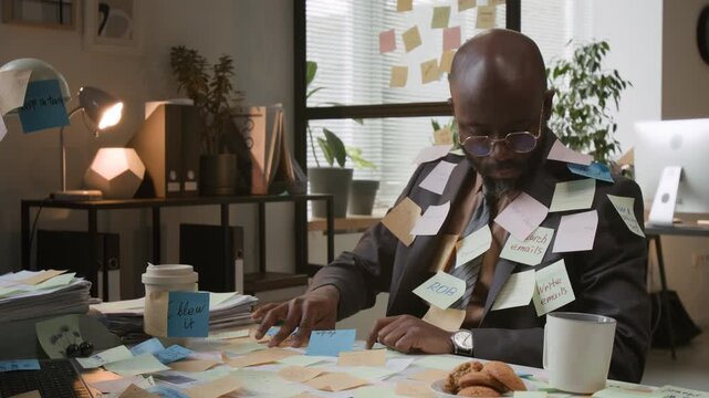 Overworked African American man, wearing glasses and business suit, covered in stickers with tasks, sitting at desk piled high with papers in office
