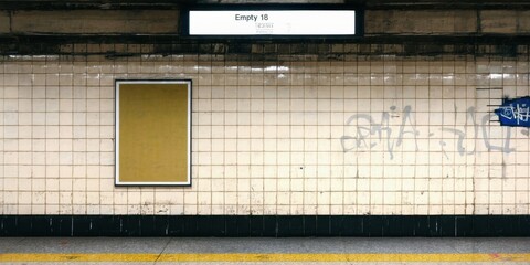 Fototapeta premium Subway station wall features blank poster space, tiled surface, graffiti. Dim lighting creates stark contrast, urban decay visible.