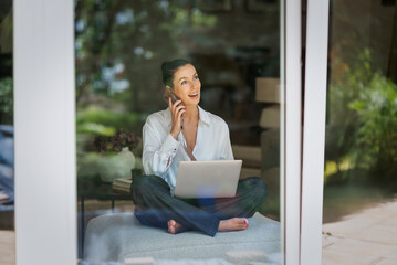 A caucasian woman photogrpahed through the window sitting on a chair and using smarpthone and laptop