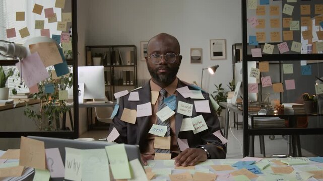Zoom out portrait shot of African American man in suit completely covered with colorful stickers with inscriptions sitting at desk littered with notes in office