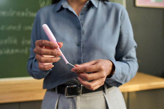 Midsection of unknown woman holding tampon in hands standing in classroom setting, demonstrating feminine hygiene product with chalkboard visible in background, face not shown