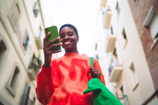 Happy African American woman looking at smartphone, mobile application, 5G data, orange sweater, green bag, authentic representation, low angle urban shot, inclusion, digital life, copy space.