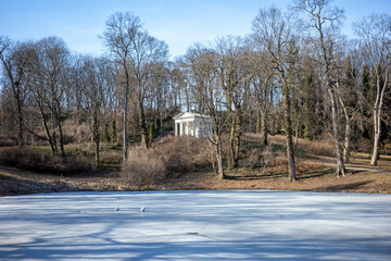 A serene winter landscape featuring a classical white pavilion nestled among bare trees by a frozen pond.