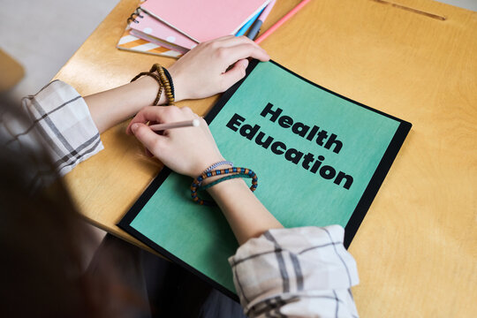 Teenager girl with light skin writing in notebook labeled Health Education, sitting at desk with hands visible, focusing on studying educational material for school assignment