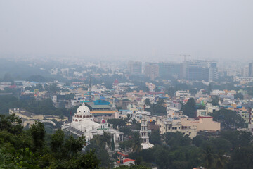 Fototapeta premium A hazy cityscape with dense buildings and a prominent church dome viewed from a green hillside in Chennai City 