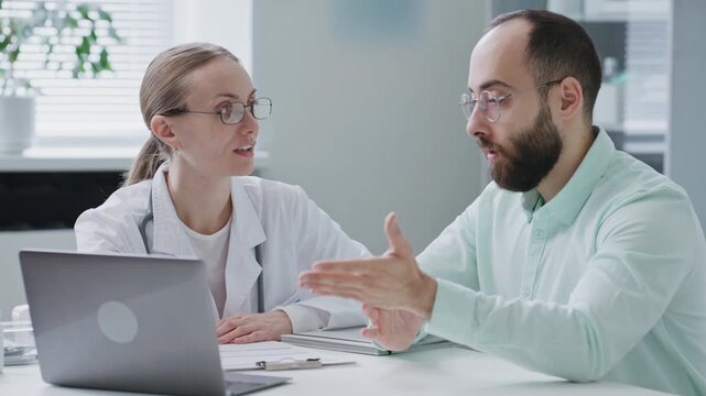 Female doctor reviewing medical information on laptop with male patient during consultation at clinic, discussing diagnosis and treatment options