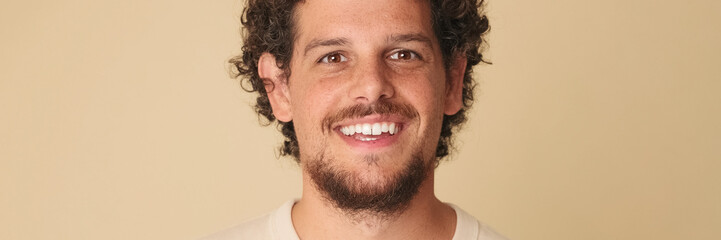 Obraz premium Close up, guy with curly hair dressed in beige t-shirt looks at the camera and laughs isolated on beige background in the studio. Panoramic