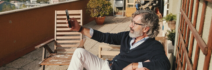 Fototapeta premium Mature man taking a selfie uses mobile phone while sitting on balcony. Panoramic